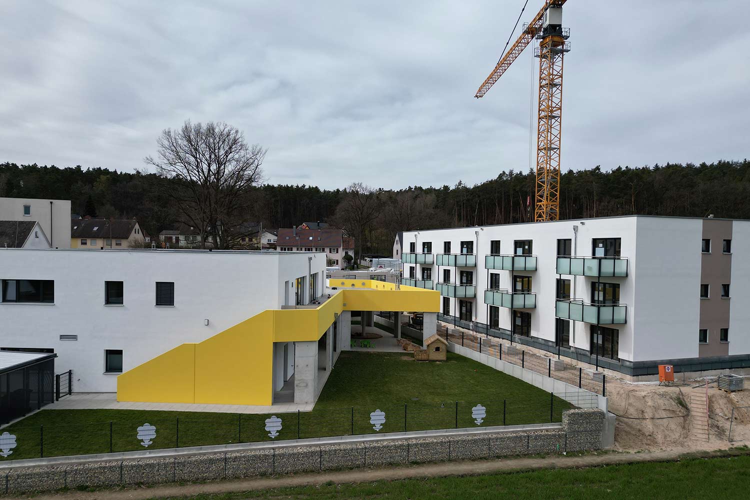 Luftaufnahme von Kita in Wolkersdorf. Daneben steht eine moderne Wohnanlage mit Flachdach.